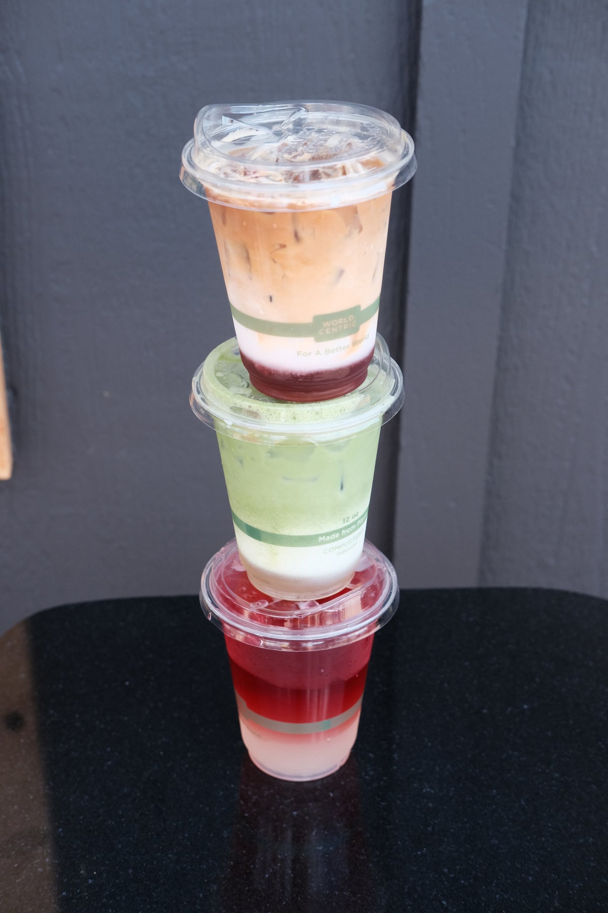Three stacked plastic cups of layered iced drinks on a black surface against a dark gray wall. The top cup is light brown and beige, the middle cup is green, white, and dark red, and the bottom cup is red, white, and pale pink.