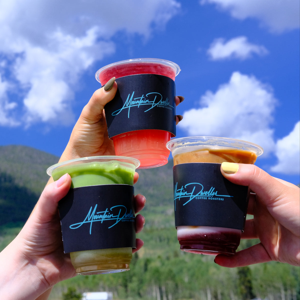 Three hands holding clear plastic cups of colorful drinks with black sleeves featuring a blue cursive logo, set against a background of green mountains and a blue sky with clouds.