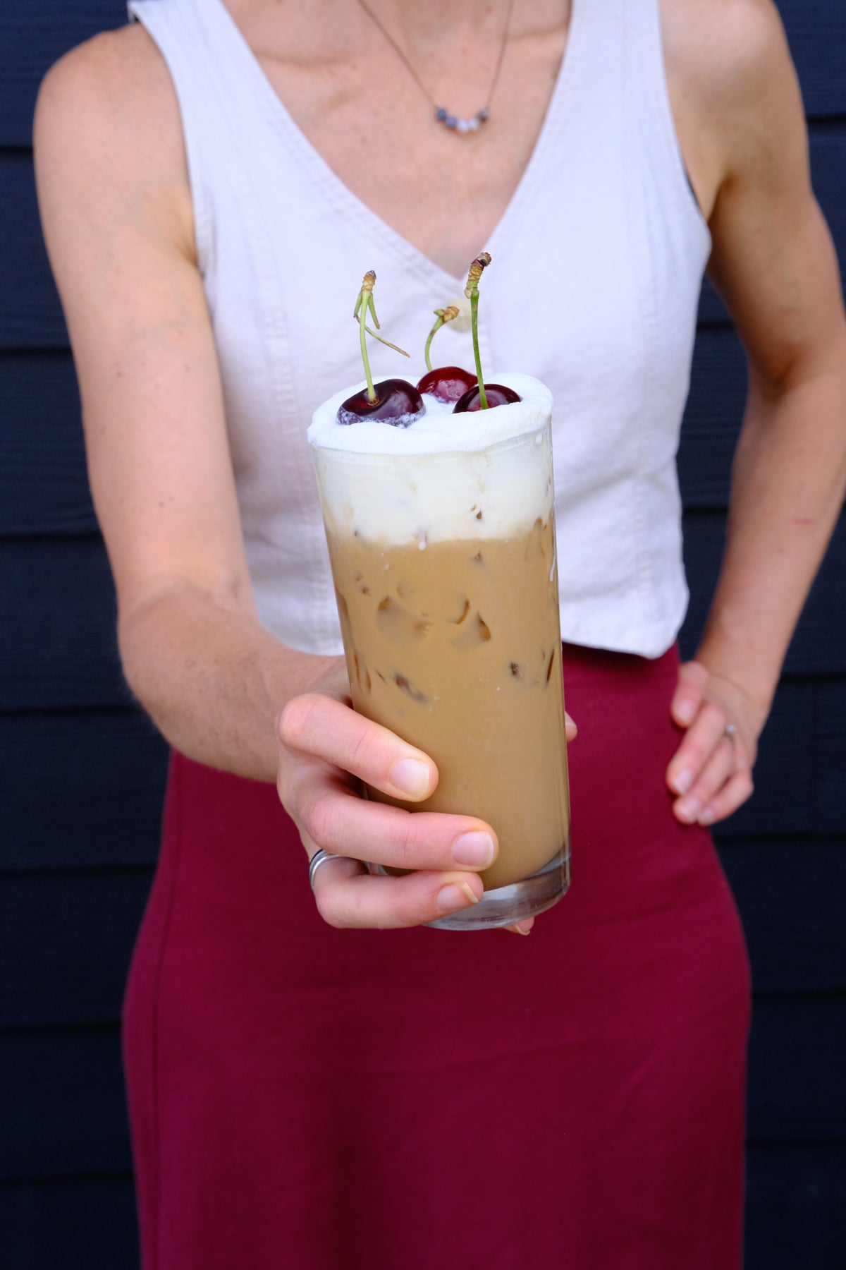 A person in a white tank top and burgundy skirt holds a tall glass of iced coffee with a thick layer of whipped cream and two cherries on top, set against a dark blue paneled wall.